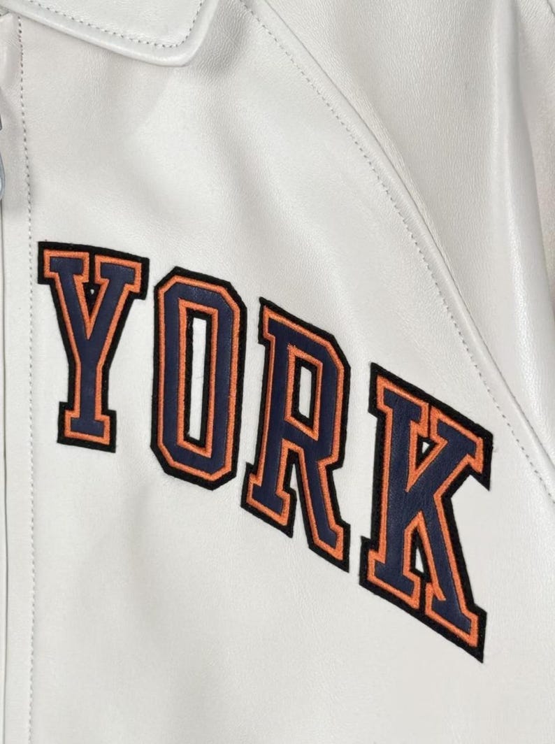 May include: Close-up of a white leather jacket with the word "YORK" embroidered on the front. The letters are navy blue with orange outlines. The jacket has a collar and a zipper.