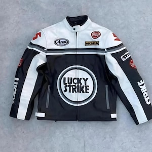 Op de afbeelding: Een zwarte, witte en grijze motorjas met de tekst "LUCKY STRIKE" op de voorkant. De jas heeft meerdere patches, waaronder "Arai" en "Michelin". De jas heeft een opstaande kraag en een drukknoopsluiting.