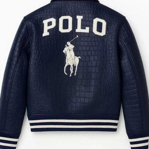 Puede incluir: Chaqueta azul marino con un patrón texturizado. La palabra "POLO" se muestra en letras blancas grandes en la parte posterior, junto con un logotipo blanco de un jugador de polo. La chaqueta tiene un ribete a rayas blanco y azul marino en la parte inferior.