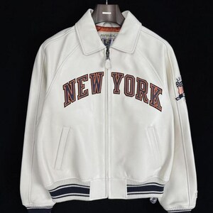 May include: Cream-colored leather jacket with the words "NEW YORK" in brown and orange lettering across the chest. The jacket has a collar, a zipper closure, and blue and white striped cuffs and hem. A small patch is on the right sleeve.