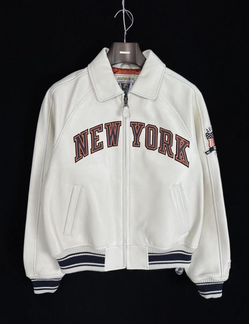 May include: Cream-colored leather jacket with the words "NEW YORK" in brown and orange lettering across the chest. The jacket has a collar, a zipper closure, and blue and white striped cuffs and hem. A small patch is on the right sleeve.