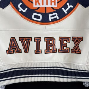 May include: Cream-colored jacket with the word "AVIREX" in orange and navy lettering. The jacket features a basketball graphic with the word "KITH" in a white box. The jacket has navy and white striped trim.