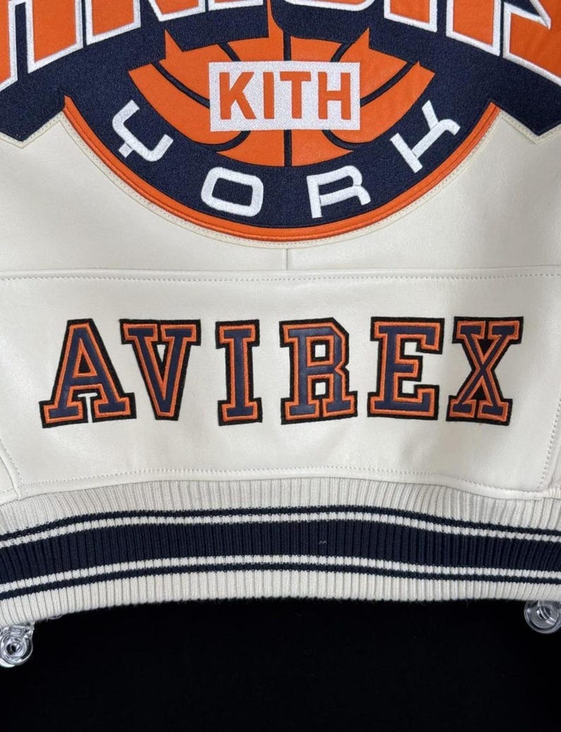 May include: Cream-colored jacket with the word "AVIREX" in orange and navy lettering. The jacket features a basketball graphic with the word "KITH" in a white box. The jacket has navy and white striped trim.