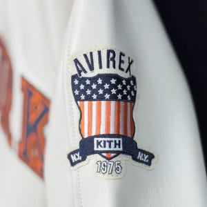 May include: Close-up of a white leather jacket featuring a patch with the "AVIREX" logo above a design of the American flag. The patch also includes "KITH" and "1975" text. The jacket has a stitched design.