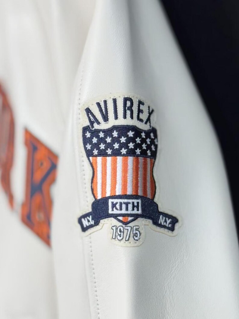 May include: Close-up of a white leather jacket featuring a patch with the "AVIREX" logo above a design of the American flag. The patch also includes "KITH" and "1975" text. The jacket has a stitched design.