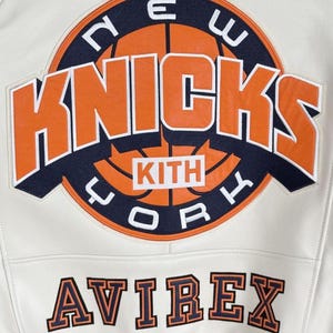 May include: Cream-colored leather jacket featuring the New York Knicks logo in orange and navy blue. The jacket includes the text "KITH" and "AVIREX" in a contrasting color. A sporty, stylish outerwear piece.