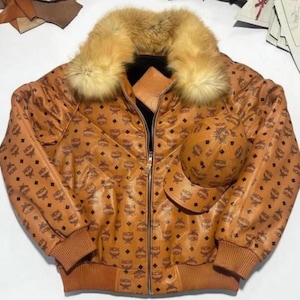 May include: A brown leather jacket and matching baseball cap set. The jacket features a fur collar and is patterned with a repeating logo design. The jacket has a zipper closure and ribbed cuffs and hem.