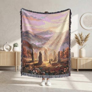 Op de afbeelding: Geweven sprei met een landschap met staande stenen, bergen en een zonsondergang. De deken heeft een gefranjerde rand en een kleurenpalet van gedempte roze, oranje en bruin. De afbeelding toont een stel dat bij de stenen staat.