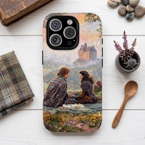 以下が含まれることがあります： 草地でブランケットに座る二人の人物のイラストがプリントされたスマートフォンケース。背景には柔らかく霞んだ空の下に石造りの城が描かれています。人物は歴史的なスタイルの衣装を身にまとっています。