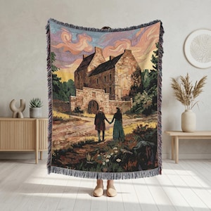 Op de afbeelding: Geweven sprei met een landschapsschilderij van een stel dat naar een kasteel loopt. Het kunstwerk toont een stenen gebouw, een pad en een kleurrijke lucht. De deken heeft een gefranjerde rand en wordt binnenshuis tentoongesteld.