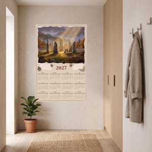 Könnte beinhalten: Ein Wandkalender für 2027 mit einer Illustration von aufrechten Steinen in einer Landschaft mit Lichtstrahlen. Der Kalender zeigt zwölf Monate in einem Raster auf hellbeigem Hintergrund mit dekorativen Elementen wie Blättern und einem Nachtfalter.