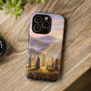 Może przedstawiać: Etui na telefon z malarstwem krajobrazu przedstawiającym parę w pobliżu kamiennych filarów. Grafika przedstawia zachód słońca nad górami i złotym polem. Etui ma błyszczące wykończenie i czarną obwódkę.