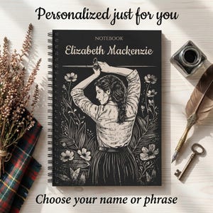 May include: Black spiral-bound notebook with the name "Elizabeth Mackenzie" on the cover. The cover features a black and white illustration of a woman holding a sword, surrounded by floral elements. Text at the top reads "Personalized just for you" and at the bottom "Choose your name or phrase."