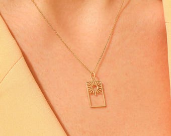 May include: A gold-colored necklace featuring a rectangular pendant with a sunburst design. The sunburst is centered within the rectangle, and the necklace is worn against a light yellow background.
