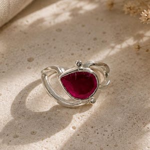 May include: A silver ring featuring a teardrop-shaped deep pink gemstone set within a curved, open-work band. The metal band has a polished finish and includes two small, clear accent stones positioned near the central pink teardrop setting.