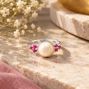 Könnte beinhalten: Ein Silberring mit einer zentralen, runden, cremefarbenen Perle in einer Fassung, die auf beiden Seiten von einer Gruppe aus drei kleinen, runden, leuchtend pinken Rubinen flankiert wird. Der Metallring besteht aus poliertem Silber.