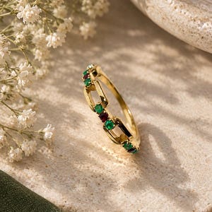 Könnte beinhalten: Ein goldener Ring mit einem Gliederkettendesign, besetzt mit abwechselnd kleinen, runden grünen und roten Edelsteinen in den Metallgliedern. Das Band hat eine polierte Oberfläche und eine feine Struktur mit einem Muster aus Steinen.