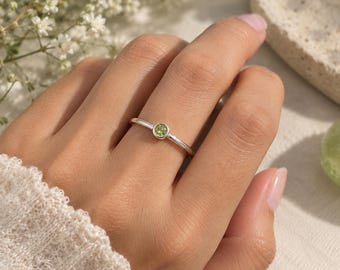 May include: A thin silver ring featuring a small, round, bezel-set light green gemstone. The band is smooth and polished, showcasing a minimalist design that centers the circular stone on the finger. The metal has a bright, reflective finish.