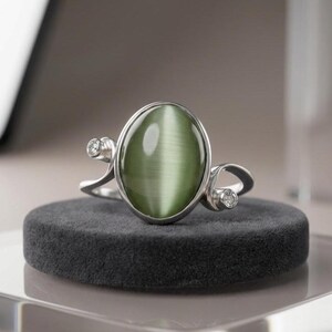 Op de afbeelding: Een zilveren ring met een grote, ovale, olijfgroene kattenoogsteen in het midden. De ringband heeft een gebogen ontwerp in bypass-stijl met een kleine, ronde, heldere edelsteen als accent aan weerszijden van de centrale zetting.