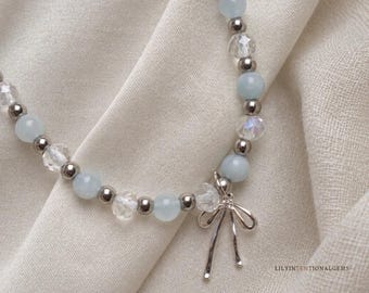Pode incluir: Um colar de contas com alternância de contas redondas azul-claras, contas transparentes facetadas e pequenas esferas metálicas prateadas. Um pingente de laço prateado pende do centro. O texto LILYINTENTIONALGEMS é visível no canto da imagem.