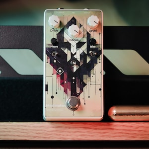 Puede incluir: Un pedal de efectos de guitarra color crema con ilustraciones geométricas negras, con perillas etiquetadas 'Drive' y 'Level'. El pedal está montado en un soporte negro con una base de madera, y un cilindro dorado descansa al lado.