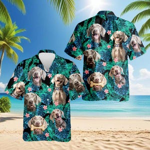 Op de afbeelding: Twee shirts met korte mouwen met een tropisch ontwerp. De shirts hebben een patroon van Weimaraner-hondengezichten, groene bladeren en roze bloemen op een zwarte achtergrond. De shirts worden op een strand getoond.