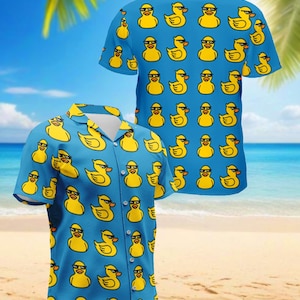 May include: Two short-sleeved button-down shirts with a repeating pattern of yellow ducks wearing sunglasses on a blue background. The shirts are displayed on a beach with palm trees and the ocean in the background.