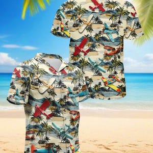 May include: Short-sleeved button-down shirt with a tropical design featuring airplanes and palm trees. The shirt has a cream background with a pattern of red, blue, and yellow airplanes, and green palm trees. The shirt is displayed on a beach setting.