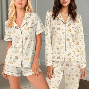 May include: Two sets of pajamas featuring a cream-colored fabric with a repeating pattern of geese, ducklings, flowers, and crescent moons. One set includes a short-sleeved button-down shirt and shorts, while the other has a long-sleeved shirt and pants.