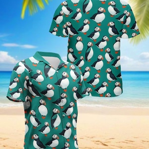 May include: A short-sleeved button-down shirt with a teal background and a repeating pattern of puffin birds. The puffins are black, white, and orange. The shirt is displayed on a beach with palm trees and the ocean in the background.
