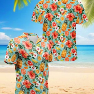Può includere: Due camicie a maniche corte con un motivo floreale e fruttato tropicale. Le camicie presentano uno sfondo azzurro con un motivo ripetuto di fiori di ibisco rossi, ananas, arance e banane. Le camicie sono esposte su una spiaggia.
