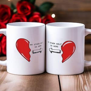 Könnte beinhalten: Zwei weiße Keramikbecher mit einem roten Herzdesign. Jeder Becher zeigt ein halbes Herz mit dem Text "MY OTHER HALF IS HERE" und einem Pfeil, der auf den anderen Becher zeigt. Die Becher stehen auf einer Holzoberfläche mit roten Rosen im Hintergrund.