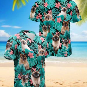 May include: A short-sleeved button-down shirt featuring a tropical design with Siamese cats, pink flowers, and green leaves on a black background. The shirt is set against a beach backdrop with palm trees and blue skies.