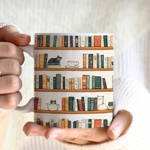 May include: White ceramic mug featuring a bookshelf design with various books and cats. The mug has a handle and is held by a person. The mug is labeled "11oz".