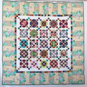 May include: A white quilt with a patchwork design featuring a grid of colorful squares. The quilt is framed by a border of floral fabric with a beige background and pink and blue flowers.