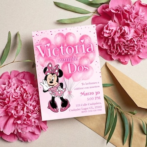 May include: Pink birthday invitation featuring Minnie Mouse holding balloons. The invitation reads "Victoria cumple Dos" with event details. Pink flowers and a brown envelope surround the invitation.