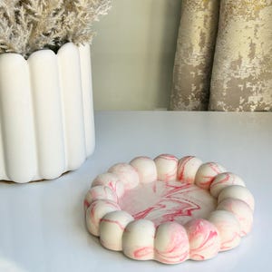 May include: A decorative tray with a unique design. The tray features a circular shape with a rim of rounded, white and pink marble-like segments. The interior of the tray has a pink and white marbled pattern. A white vase with dried plants is in the background.