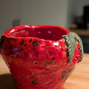 Könnte beinhalten: Handgefertigte Keramikschale in Erdbeerform. Die Schale ist leuchtend rot mit dunkelgrünen Sprenkeln und einem grünen Blattdetail. Der Innenraum ist glänzend rot.