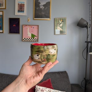Może przedstawiać: Ceramiczna miska z żywym czerwonym wnętrzem i zieloną stroną zewnętrzną, ozdobioną ręcznie malowanymi białymi owcami pasącymi się na polu. Miska ma zaokrąglony kształt i gładkie wykończenie, przedstawiające szczegółową scenę pasterską.