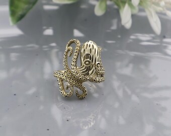 Peut inclure: Une bague en forme de pieuvre de couleur or. La bague présente un design détaillé d'une pieuvre avec des tentacules, une tête texturée et des yeux circulaires. La bague est posée sur un fond doux et neutre, mettant en valeur les détails.