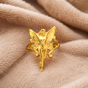 May include: A gold-toned ring featuring a central fairy figure with outstretched wings. The fairy has a slender form and detailed wing patterns. The band of the ring extends from the sides of the fairy figure, creating a cohesive and singular piece of jewelry.