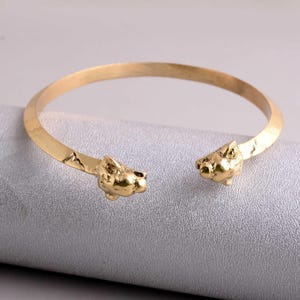 Puede incluir: Brazalete dorado con esculturas de cabezas de animales en los extremos. El brazalete abierto presenta cabezas de animales detalladas. Hecho de metal brillante. Circunferencia de aproximadamente 17 cm.