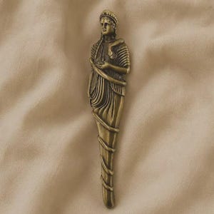 Pode incluir: Um objeto decorativo em metal de tom bronze com a forma de uma figura em pé vestida com túnicas fluidas. A figura usa uma faixa na cabeça e é entrelaçada por uma cobra longa e fina que se enrola no tronco e nas pernas até à base.