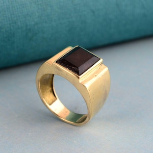 Pode incluir: Um anel dourado com uma pedra escura de corte quadrado. O anel tem uma banda grossa e uma moldura retangular ao redor da pedra. O anel é exibido em um fundo azul-petróleo.