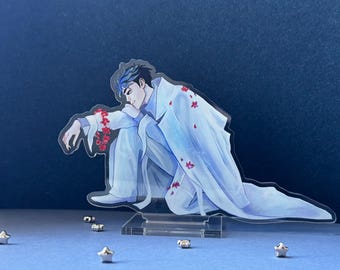 May include: A clear acrylic standee featuring an illustrated character with dark hair and blue highlights, wearing a long white coat and white trousers. The character is in a crouching pose, adorned with red floral accents on the arm and coat.