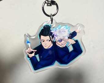 May include: A clear acrylic keychain featuring two anime characters in dark blue outfits. One character has black hair and a blue flame, the other has white hair and sunglasses. The keychain has a silver metal clasp.