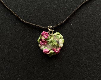May include: A heart-shaped pendant necklace with a dark brown cord. The pendant is adorned with pink and white flowers and green leaves. The pendant is attached to a gold-colored clasp.