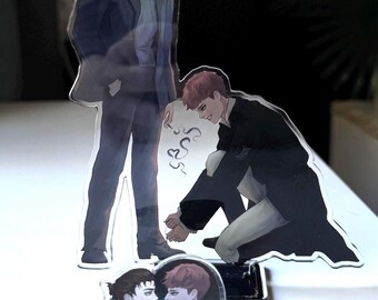May include: Acrylic standees featuring anime-style illustrations of two male figures. One stands, the other kneels, with a heart-shaped pin featuring the same characters. The figures are in shades of black, blue, and gray.