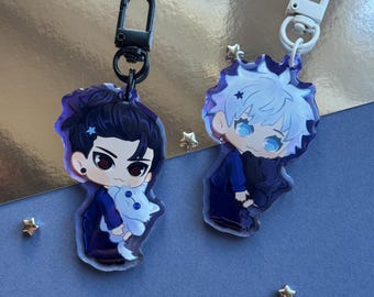 May include: Two acrylic keychains featuring chibi-style anime characters. One keychain has a black clasp and a character with dark hair holding a white creature. The other has a white clasp and a character with white hair holding a dark creature.