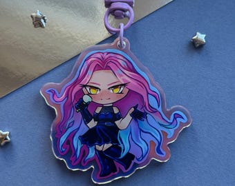 May include: A clear acrylic keychain featuring a chibi-style character with long, flowing pink and blue hair, yellow eyes, and a blush on their cheeks. The character wears a dark blue dress and gloves while holding a microphone, attached to a light purple clasp.